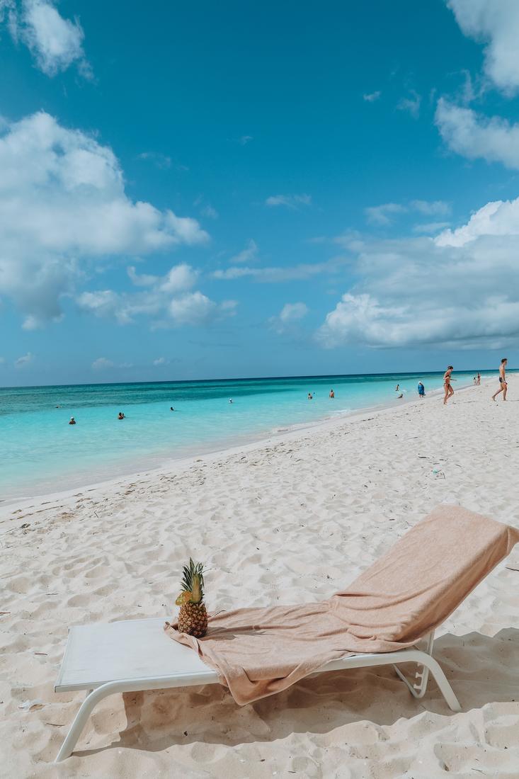 A beach lounger with a pineapple drink on white sand beside turquoise water