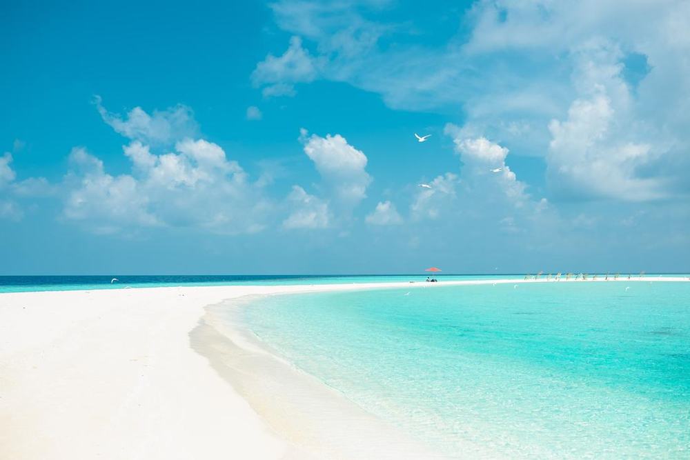 A curved white sand bar and turquoise shallows with a red umbrella in the distance