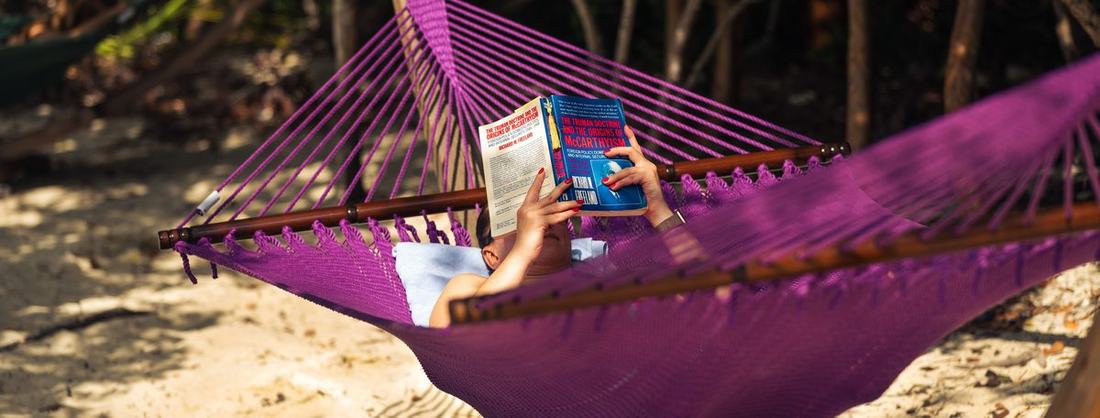 A woman reading in a hammock in dappled shade on a sandy beach