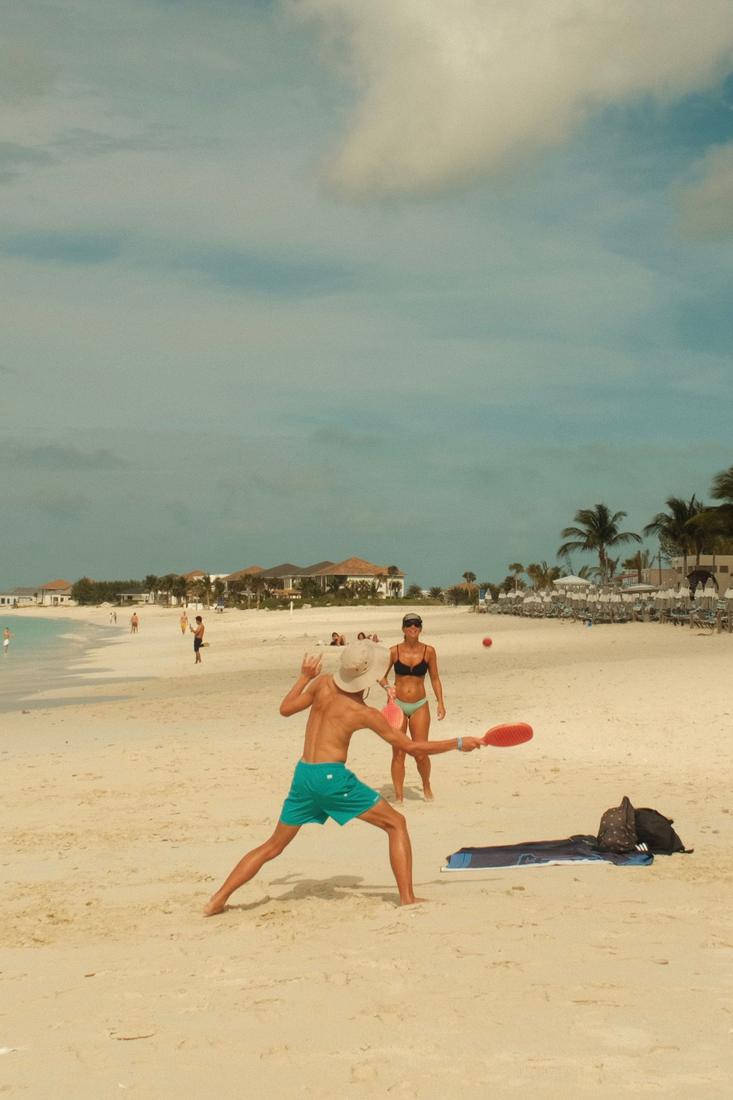 Two people playing beach paddle ball on a sandy beach with homes in the background